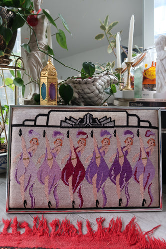 Decorative needlepoint with burlesque dancers with a silver frame, surrounded by plants and decor items.