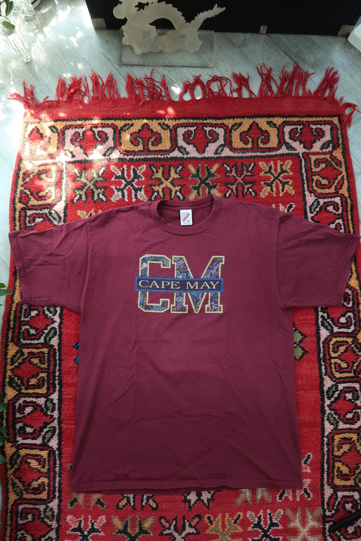 Maroon t-shirt with 'CM' logo on a red patterned rug