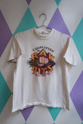 A white vintage graphic t-shirt hanging on a metal hanger against a purple, teal, and white diamond-patterned wall. The graphic features a fist crushing wheat with the text 'I QUALIFIED FOR RAW BITS'.