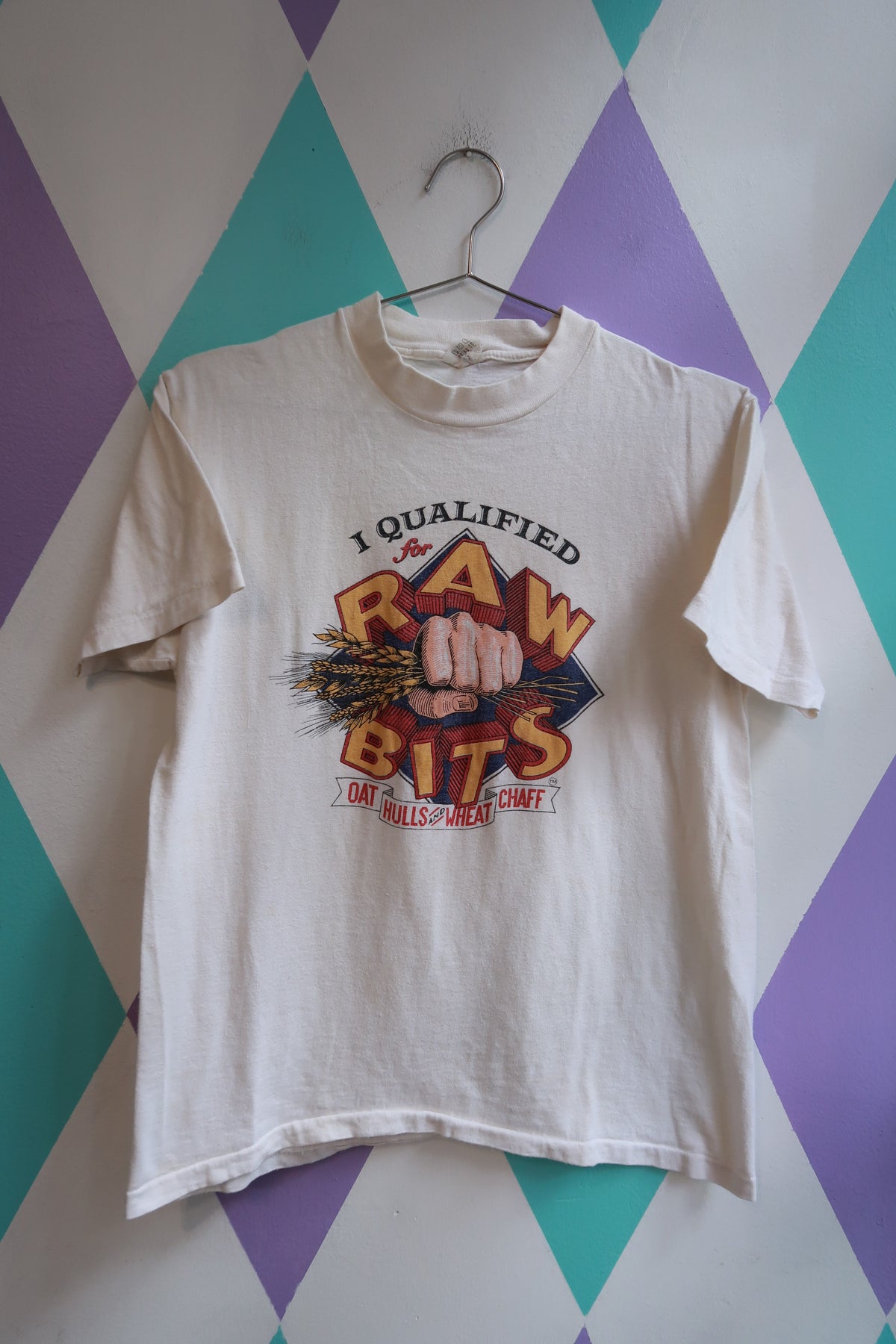 A white vintage graphic t-shirt hanging on a metal hanger against a purple, teal, and white diamond-patterned wall. The graphic features a fist crushing wheat with the text 'I QUALIFIED FOR RAW BITS'.
